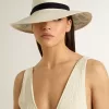 Gouden fedora hoed van wit stro met zwart lint en maritieme print onder de rand.