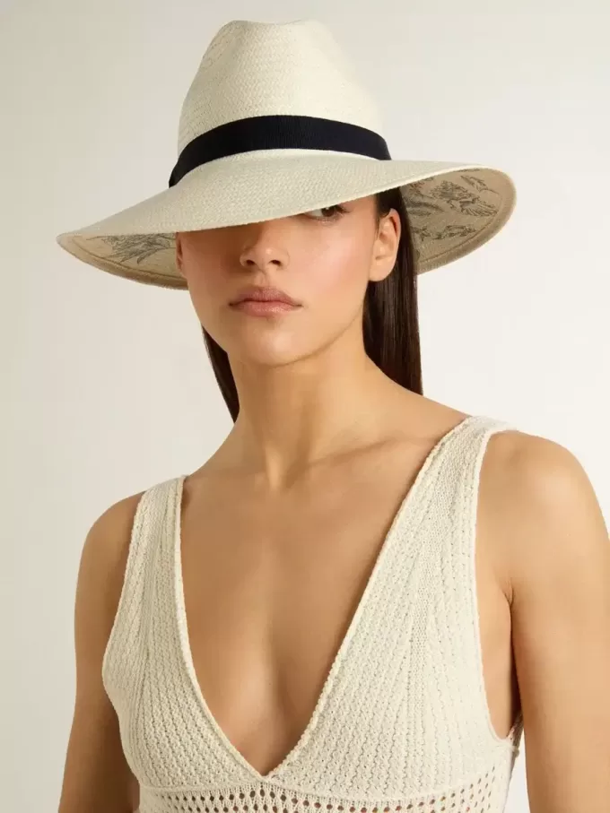 Gouden fedora hoed van wit stro met zwart lint en maritieme print onder de rand.